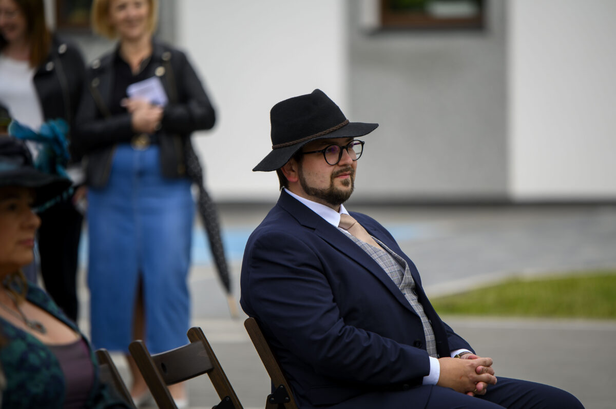 Mężczyzna w ciemnym garniturze i kapeluszu siedzi na zewnątrz. Za nim stoją osoby w jasnym tle budynku. Atmosfera formalna.