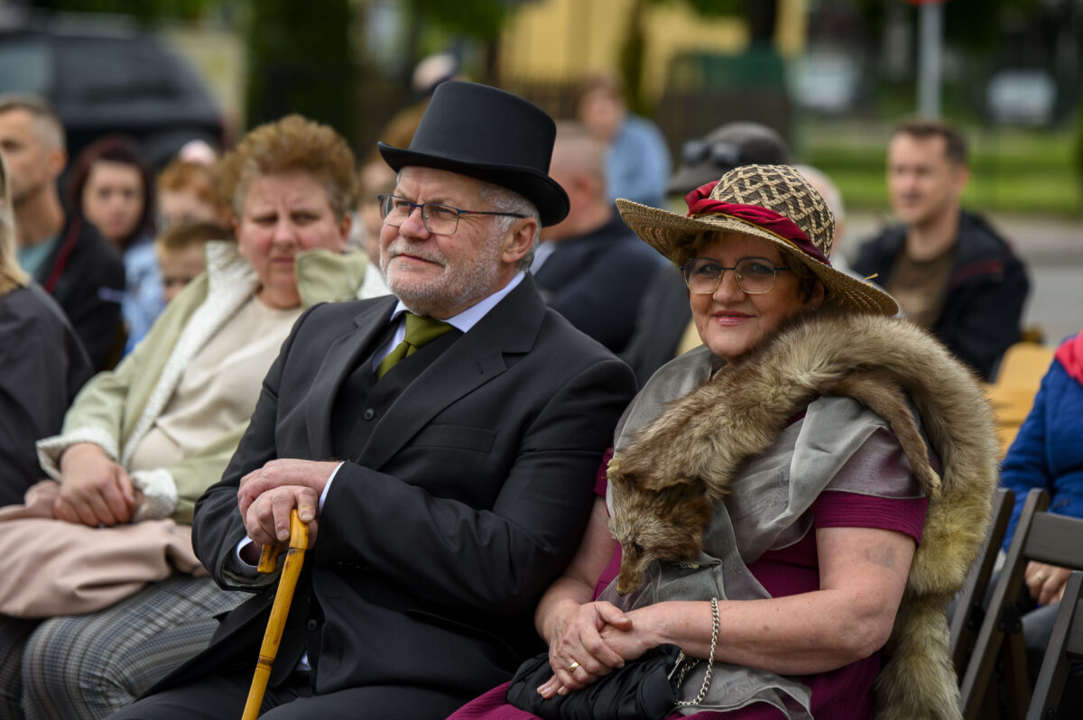 Para w eleganckich, retro strojach siedzi na zewnątrz. Mężczyzna ma czarny frak i cylinder. Kobieta w kapeluszu i futrze, z uśmiechem.