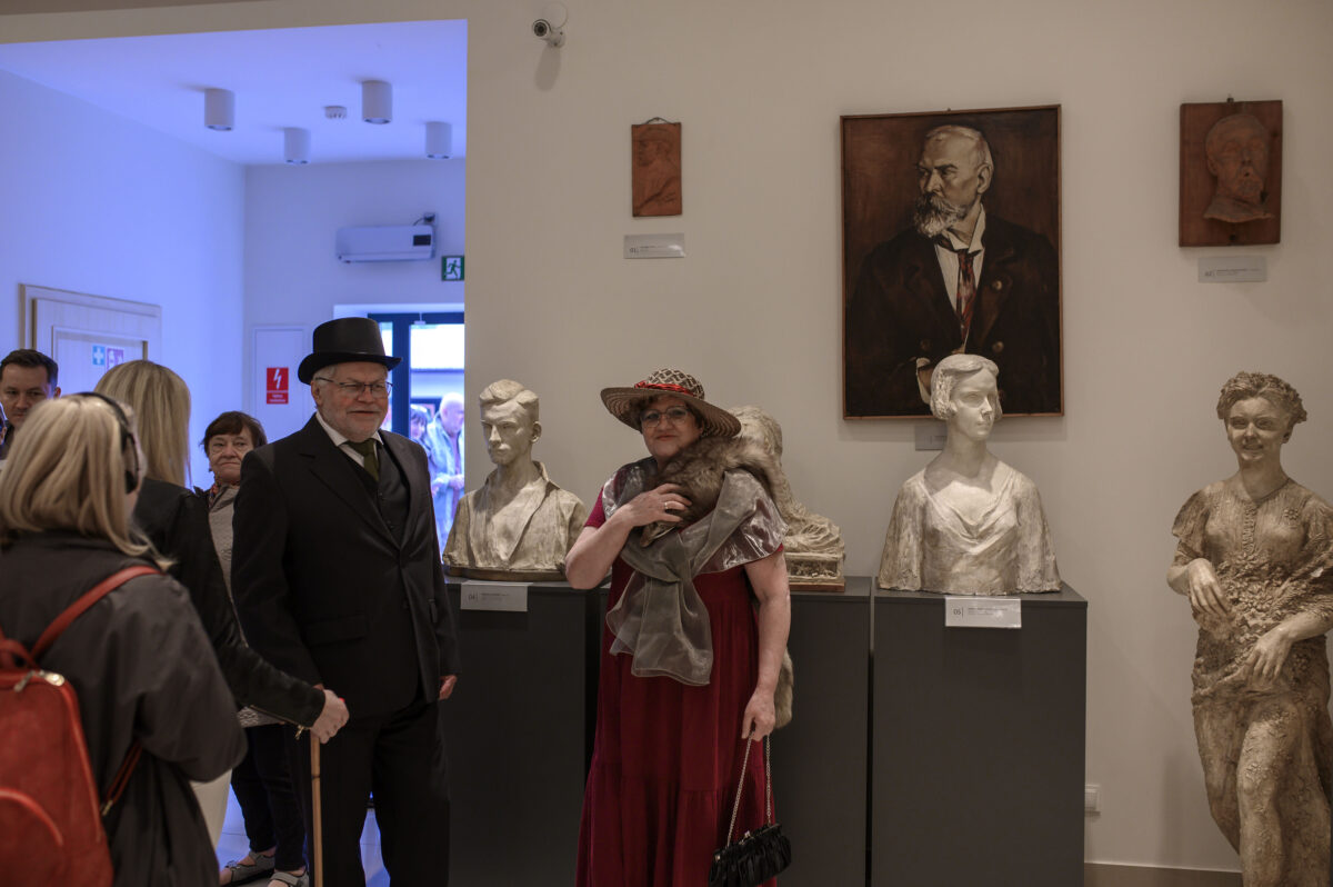 W muzeum, ludzie w strojach z epoki i rzeźby na postumentach. Dominuje biel ścian i kilka dzieł sztuki na tle. Atmosfera retro w eleganckim otoczeniu.