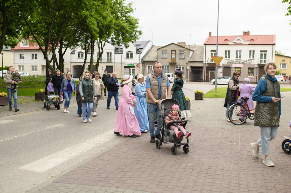 Grupa osób spaceruje po ulicy obok kamienic, część ubrana w historyczne stroje. Przewaga zieleni i pastelowych barw. Na trotuarze wózek z dzieckiem.