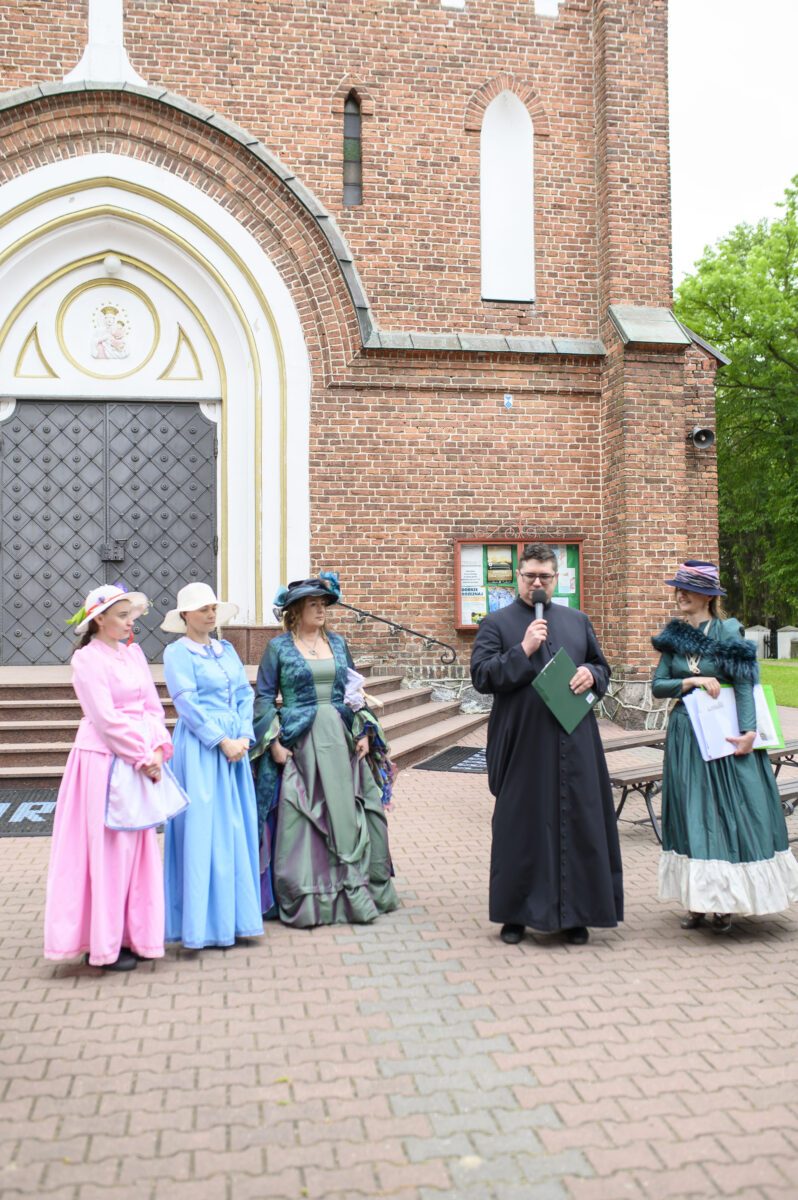 Grupa osób w kolorowych strojach historycznych stoi przed ceglaną fasadą kościoła. Na środku mężczyzna w czarnym stroju, trzyma mikrofon. Tło stanowi drzwi kościoła i zieleń po prawej stronie.