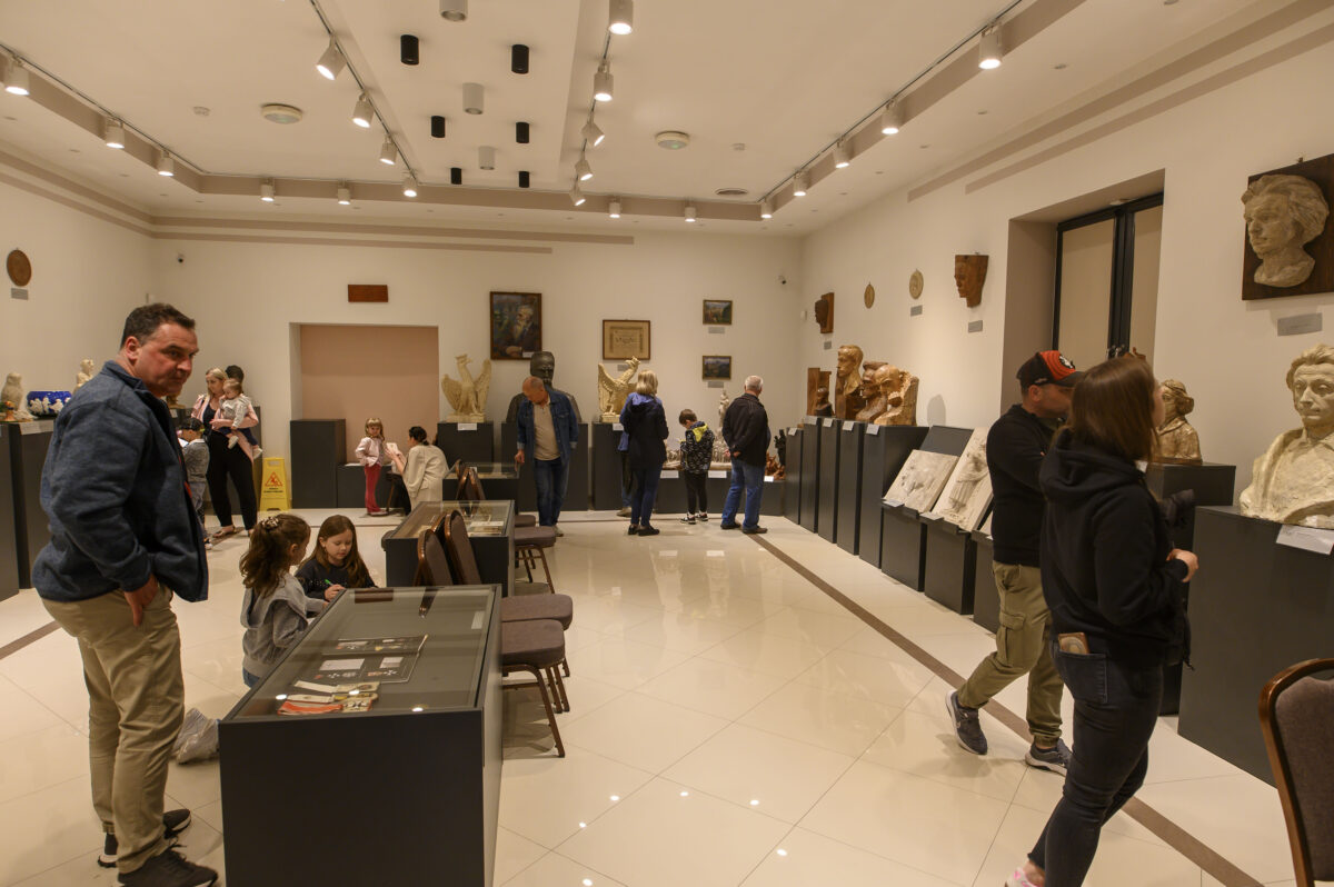 Wnętrze muzeum z rzeźbami na ciemnych podestach. Kilka osób ogląda eksponaty w jasnym pomieszczeniu. Sufit ozdobiony reflektorami.