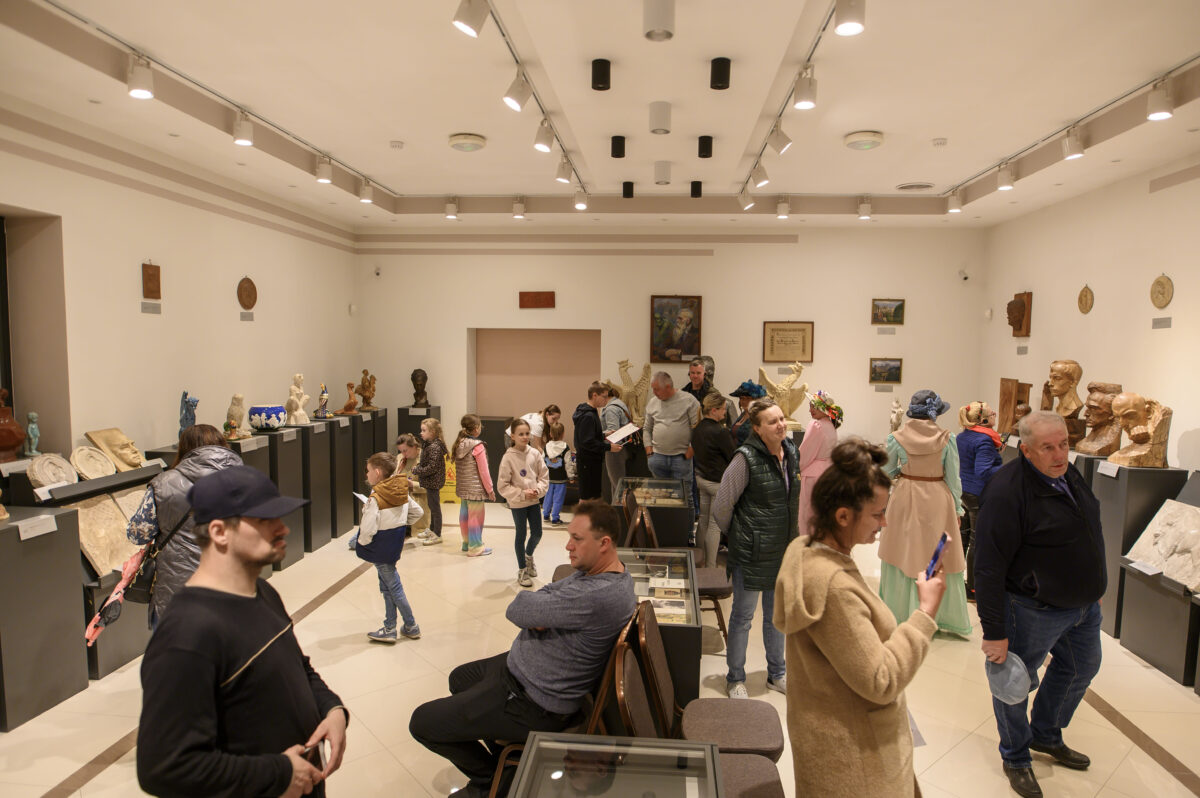 Sala galerii z białymi ścianami, rzeźbami na podestach, pełna zwiedzających. Jasne światło podkreśla detale eksponatów. Tło neutralne, podłoga w odcieniach beżu.
