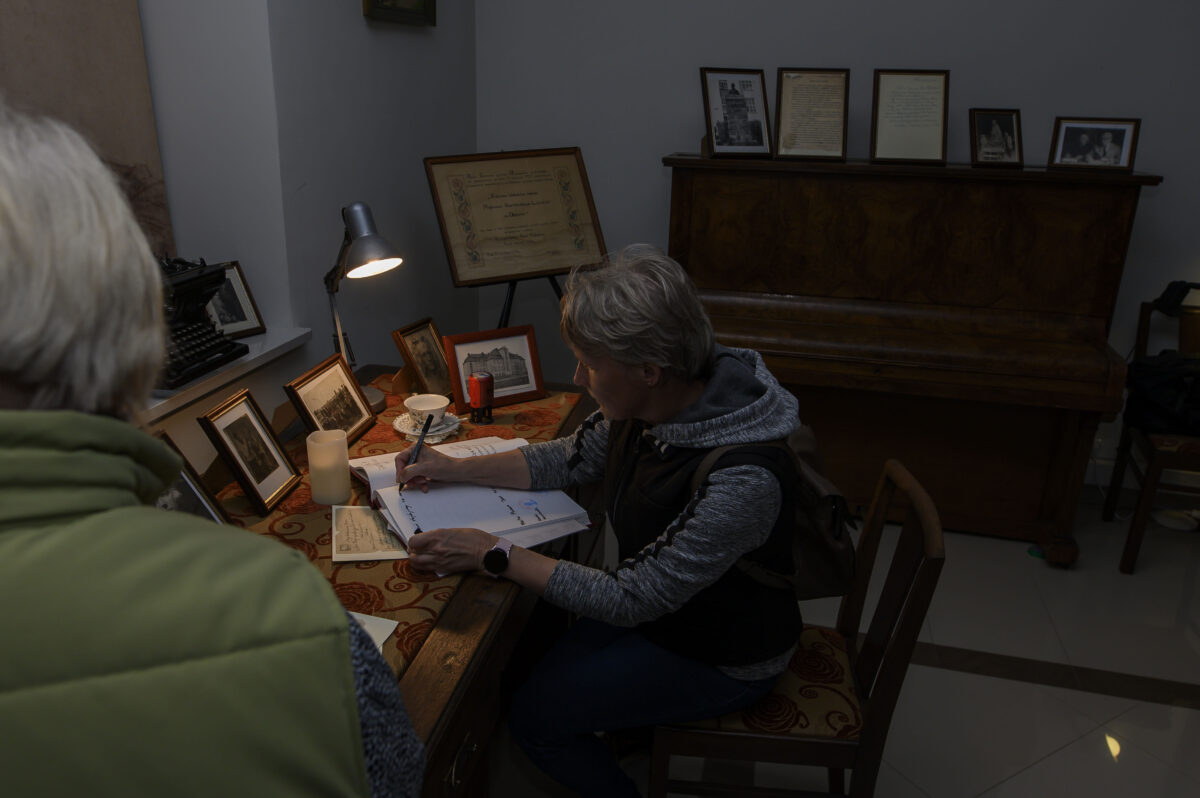 Osoba przy biurku pisząca w notatniku, otoczona starymi zdjęciami. Światło lampki biurkowej oświetla przestrzeń. W tle brązowe pianino z ramkami.