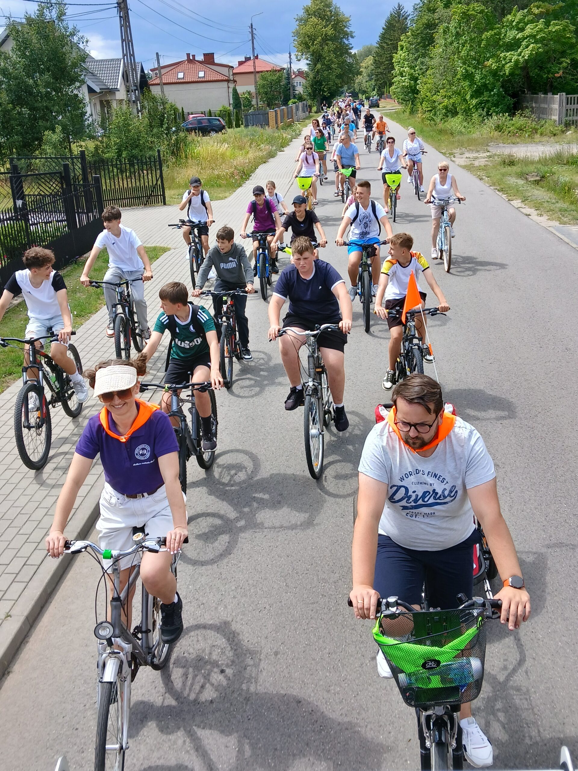 Grupa rowerzystów jedzie ulicą w słoneczny dzień. Widoczne zielone krzewy i domy w tle. Różnorodne stroje, kaski i plecaki.