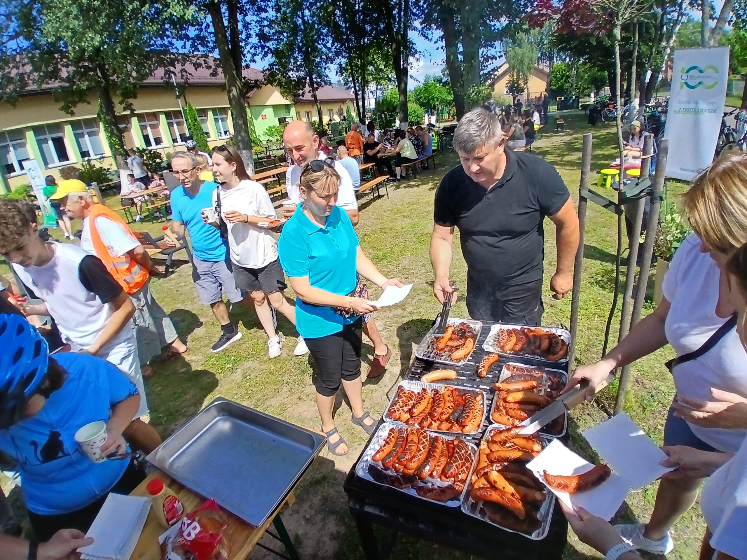 Grupa ludzi na pikniku próbuje kiełbasek z grilla. Tło stanowi zielony teren z drzewami i budynkami. Pogoda jest słoneczna, co podkreślają jasne kolory ubrań uczestników.