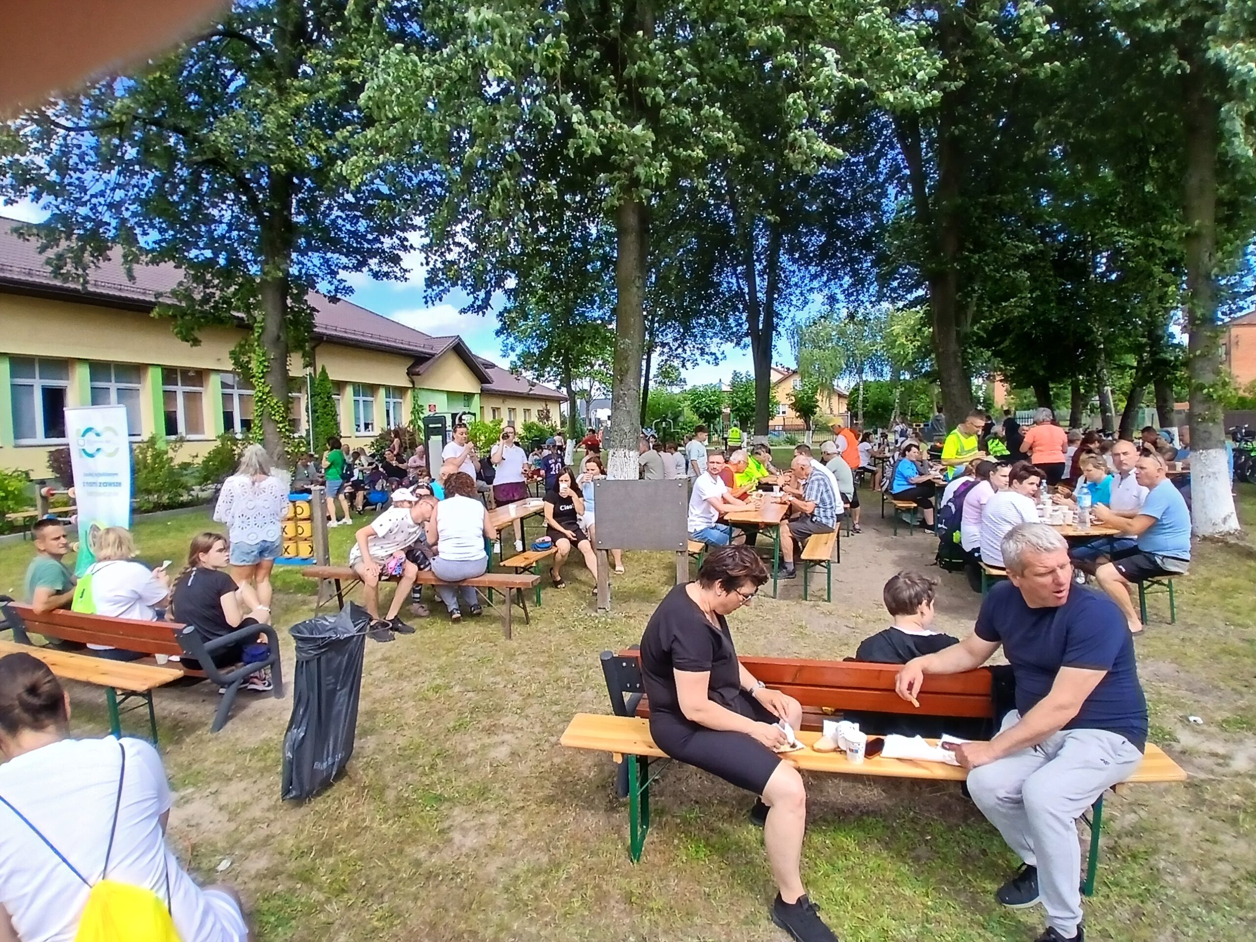Na zdjęciu widać ludzi siedzących na ławkach pod drzewami na pikniku. Tło stanowi budynek z kremową elewacją. Dominuje zielona i niebieska kolorystyka.