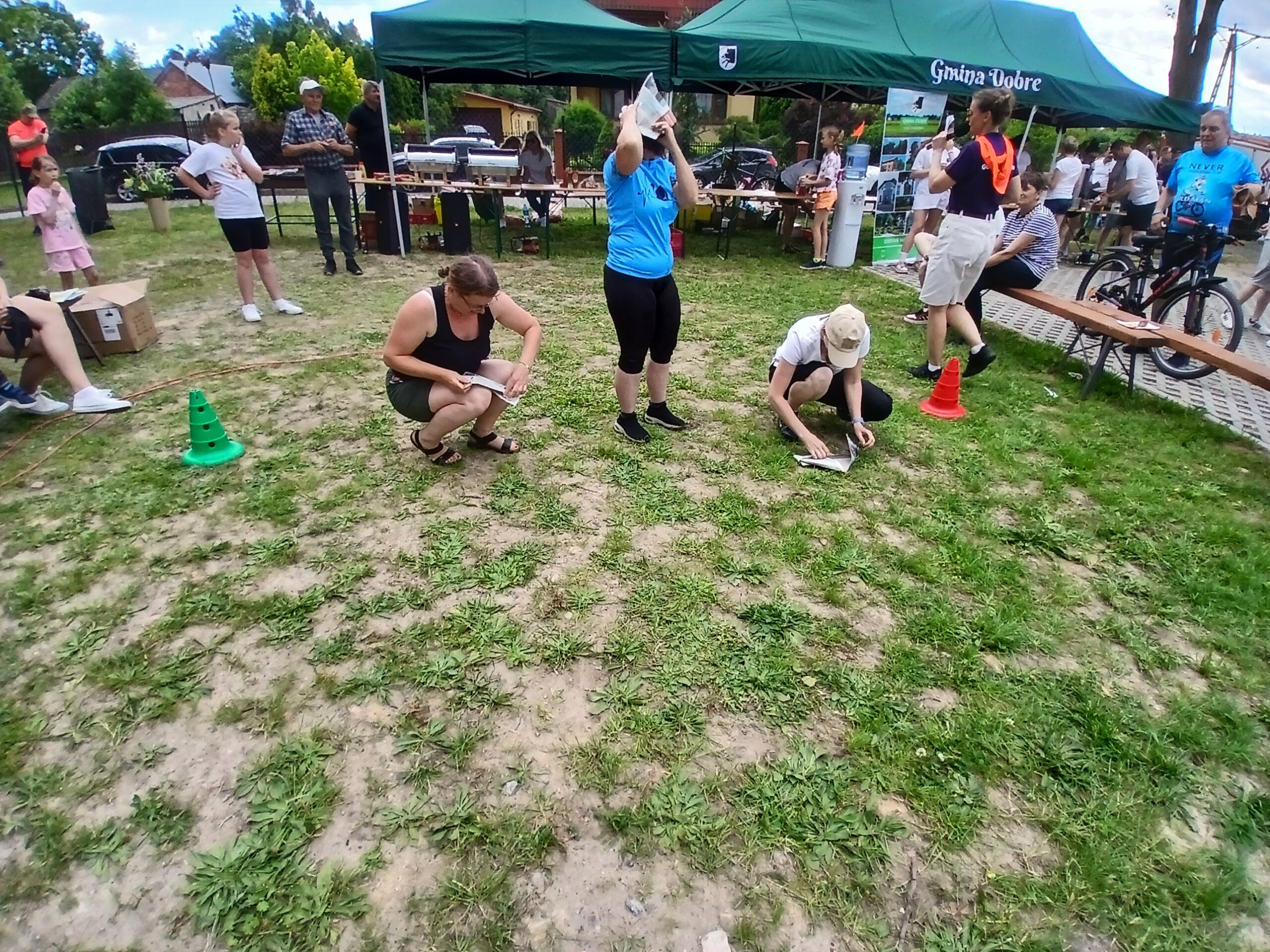 Grupa ludzi na trawie, biorących udział w aktywności. Zielony namiot w tle, kolorowe stożki na ziemi. Dzień słoneczny, atmosfera piknikowa.