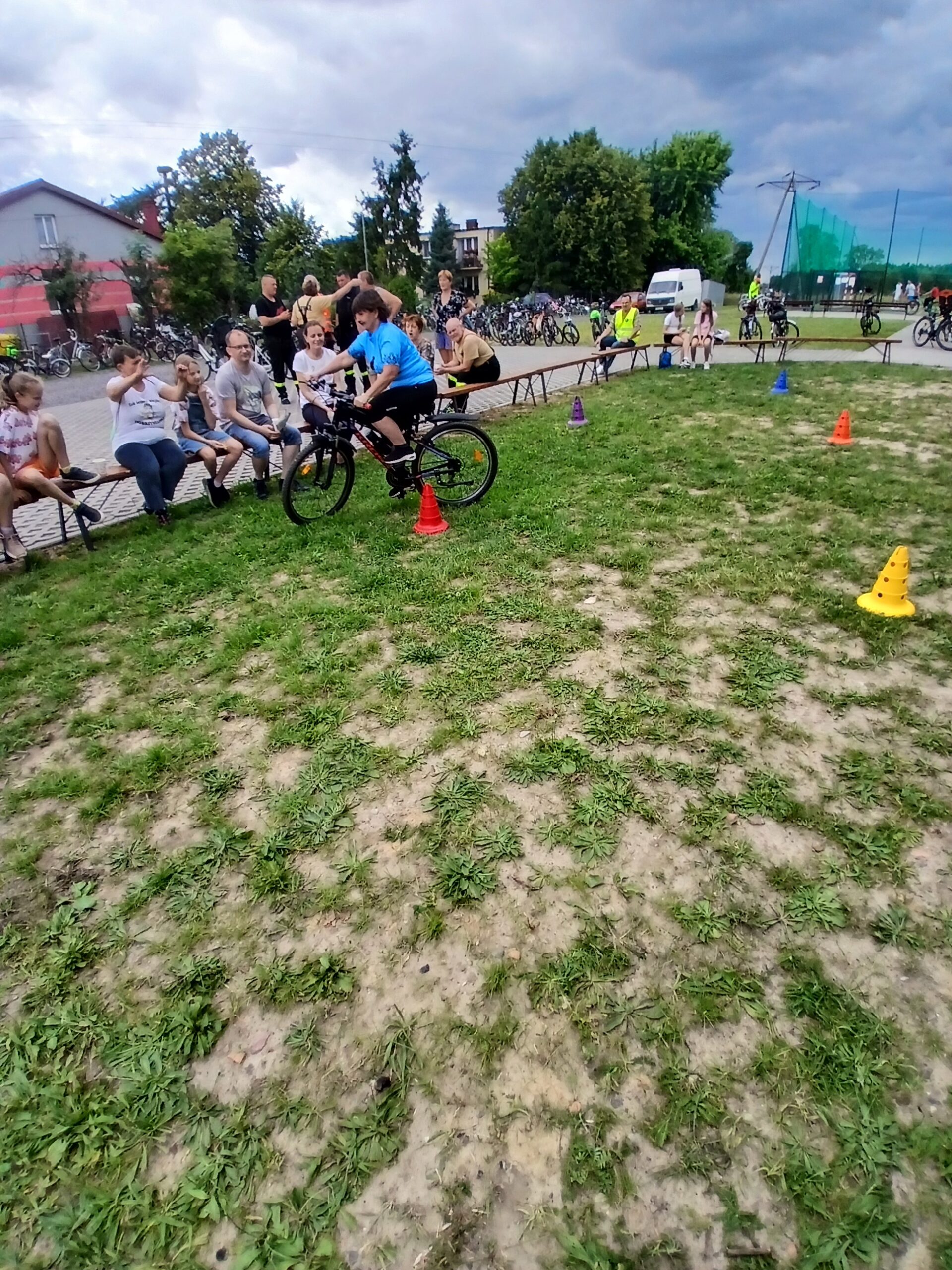 Na zdjęciu widać osobę na rowerze, manewrującą między kolorowymi pachołkami na trawie. W tle siedzą ludzie na ławkach, niebo jest pochmurne. Otoczenie jest zielone z kilkoma drzewami.