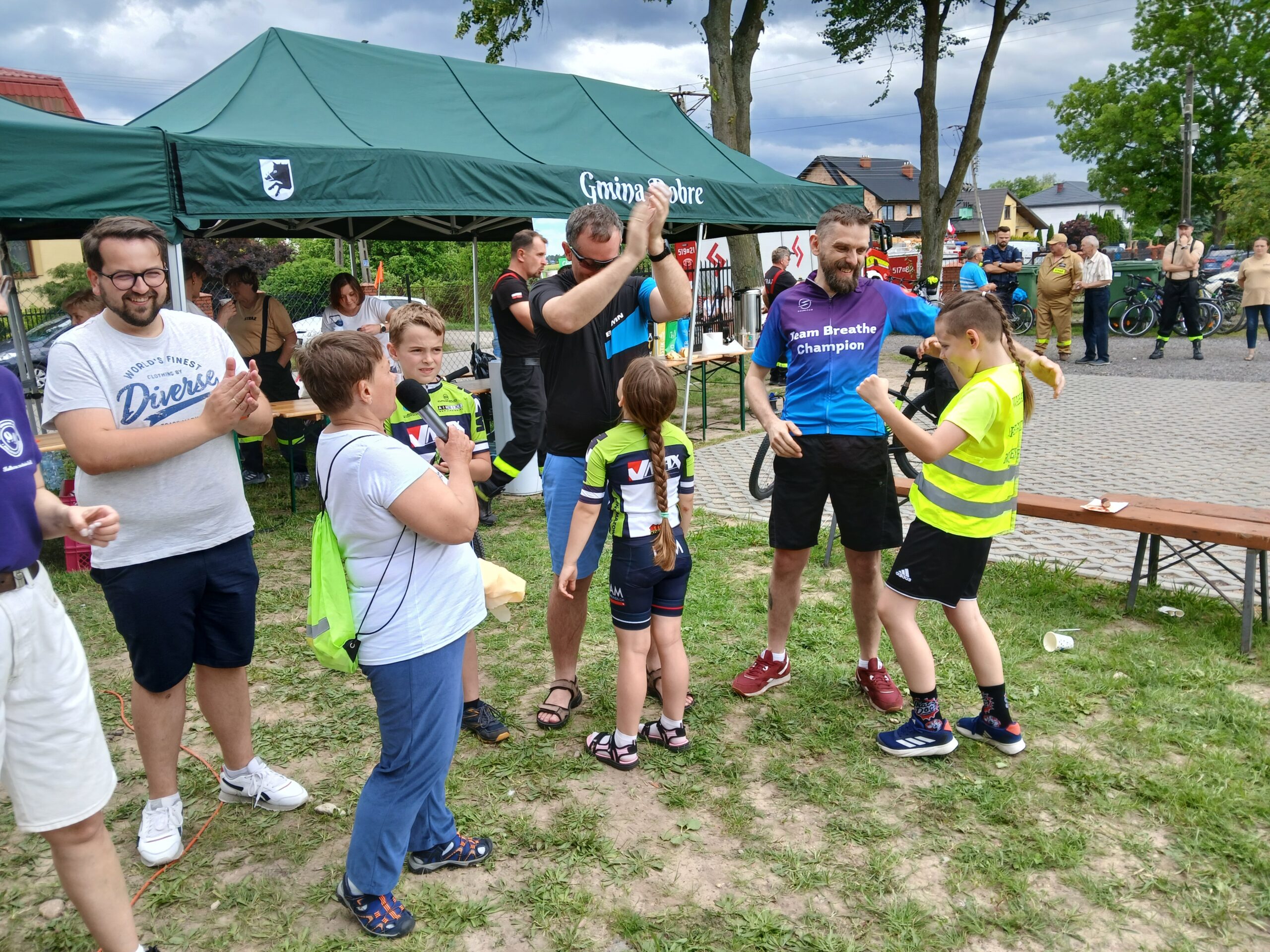 Grupa ludzi stoi na trawie, obok zielonego namiotu. Kilka osób w sportowych ubraniach, w tle są drzewa i budynki. Atmosfera przypomina festyn lub wydarzenie sportowe.