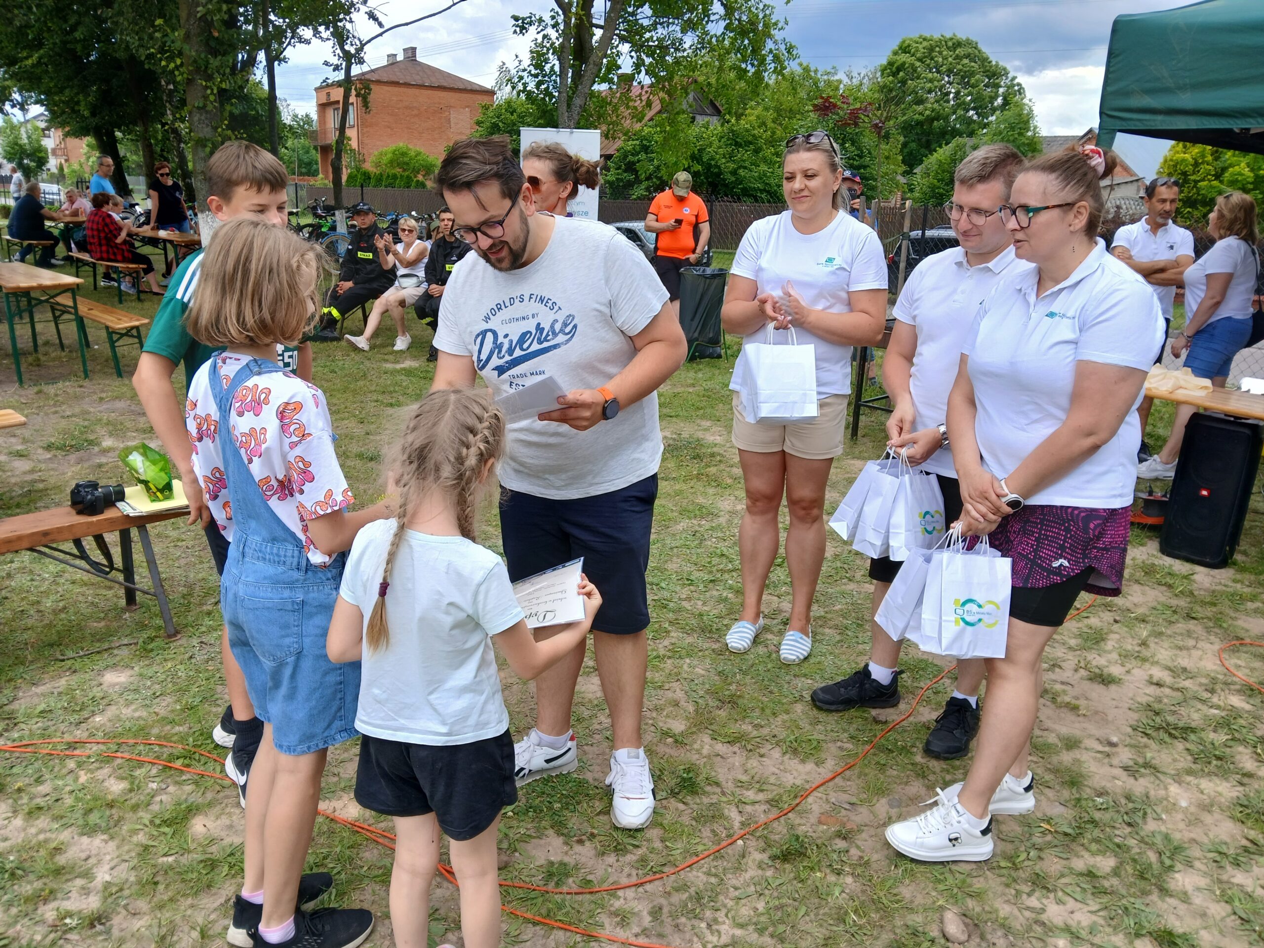 Grupa ludzi na pikniku, dzieci otrzymują torby z upominkami. Trawa i drzewa w tle, niebieskie i białe ubrania dominują.