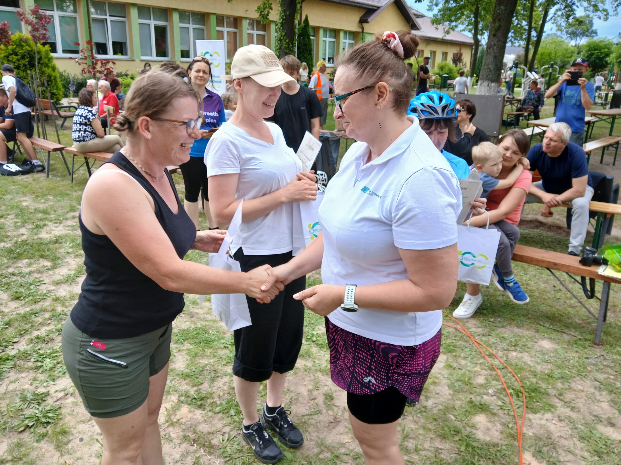 Grupa ludzi na zewnątrz, kobiety podają sobie ręce. Zielona trawa, stoliki piknikowe w tle. Relaksująca, przyjacielska atmosfera.
