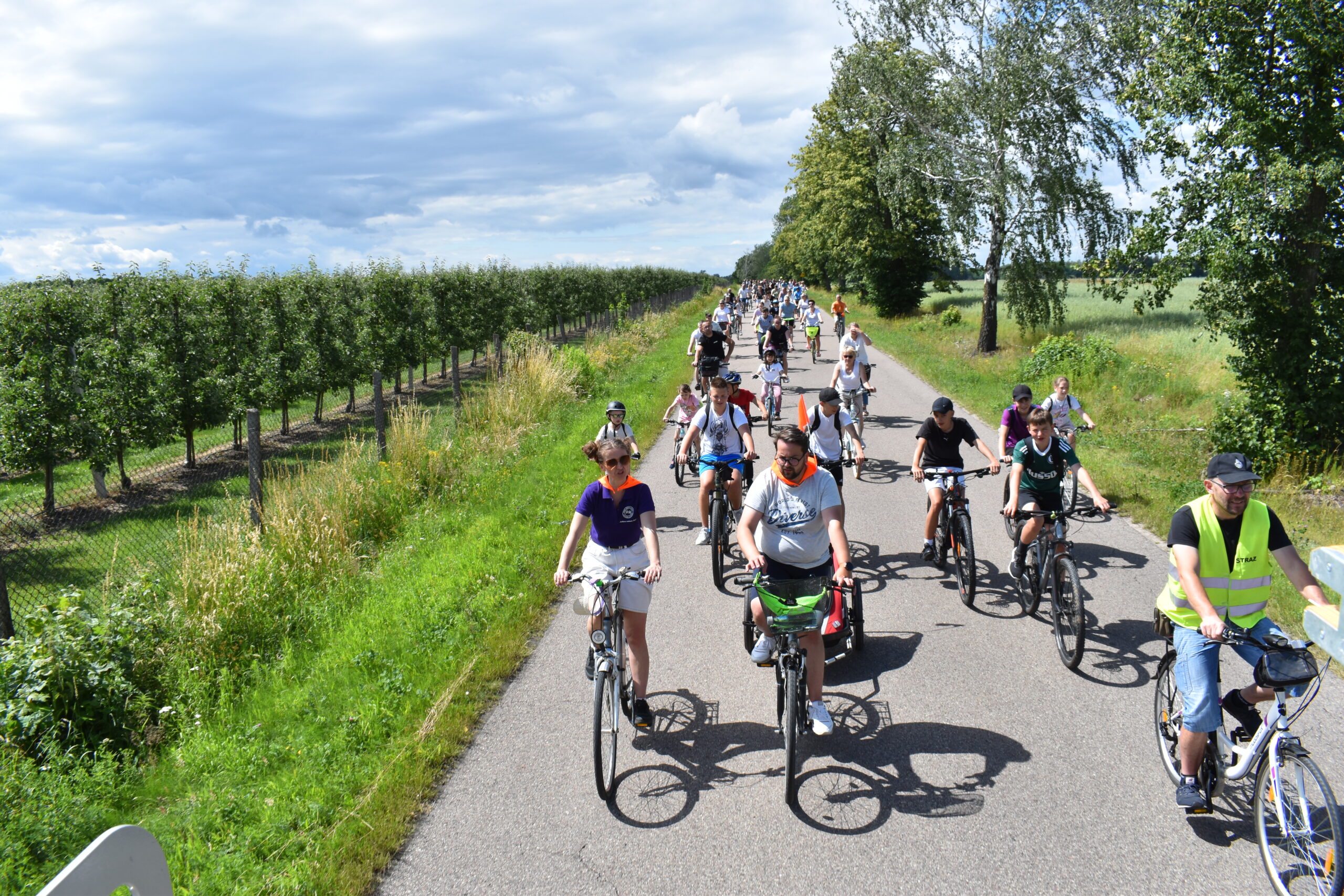Grupa rowerzystów jedzie asfaltową drogą wśród zieleni. Po lewej stronie widać rząd drzew owocowych. Pogoda jest słoneczna z niewielkim zachmurzeniem.