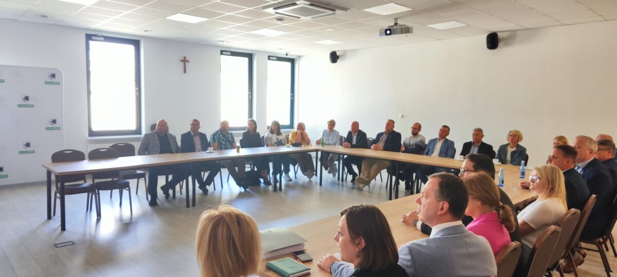 Zdjęcie przedstawia grupę ludzi siedzących przy dużym prostokątnym stole w jasnym pomieszczeniu. Na ścianie w tle widać krzyż i okna z jasnym światłem. Osoby noszą formalne ubrania, a oczy są zwrócone w jednym kierunku.