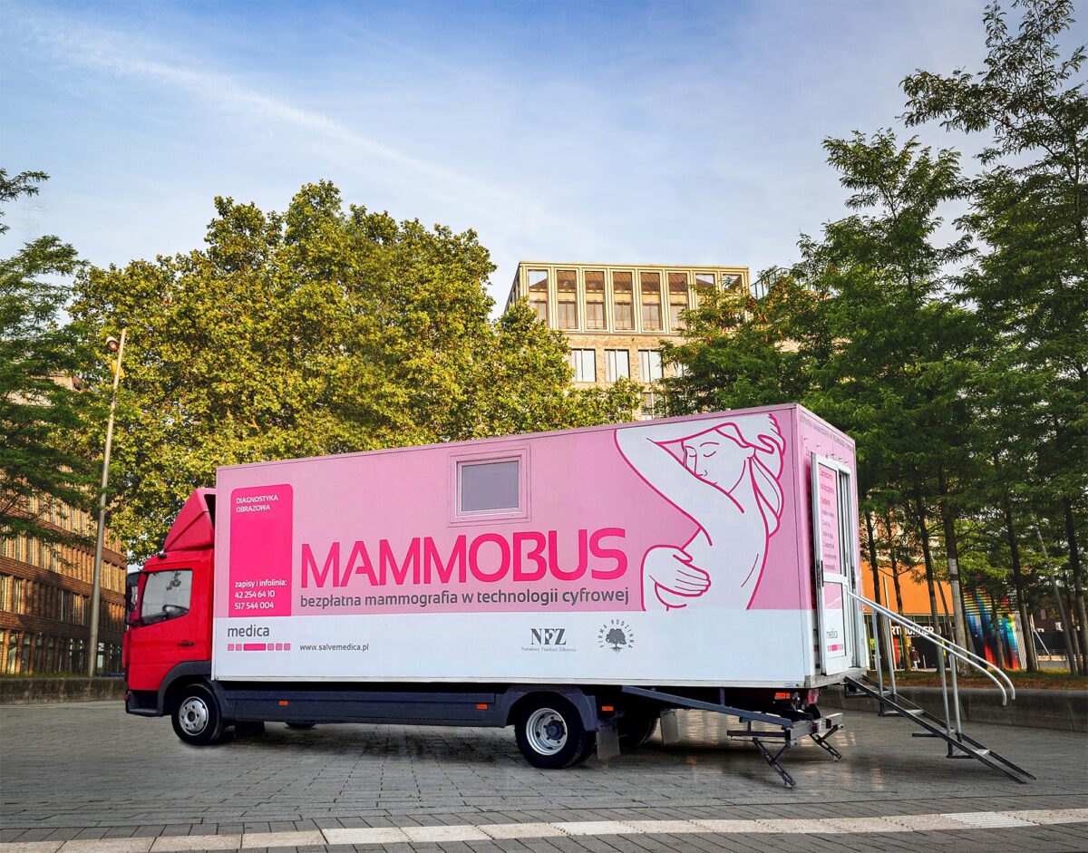 Czerwony mammo-bus z różowym napisem stoi na placu. W tle drzewa, budynek i jasne niebo. Oferuje darmową mammografię.