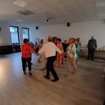 Grupa starszych osób tańczy w jasnym pomieszczeniu z drewnianą podłogą. Ściany są białe, a na nich okna z niebieskim światłem. Ubrania mają różne kolory, m.in. czerwień i zieleń.