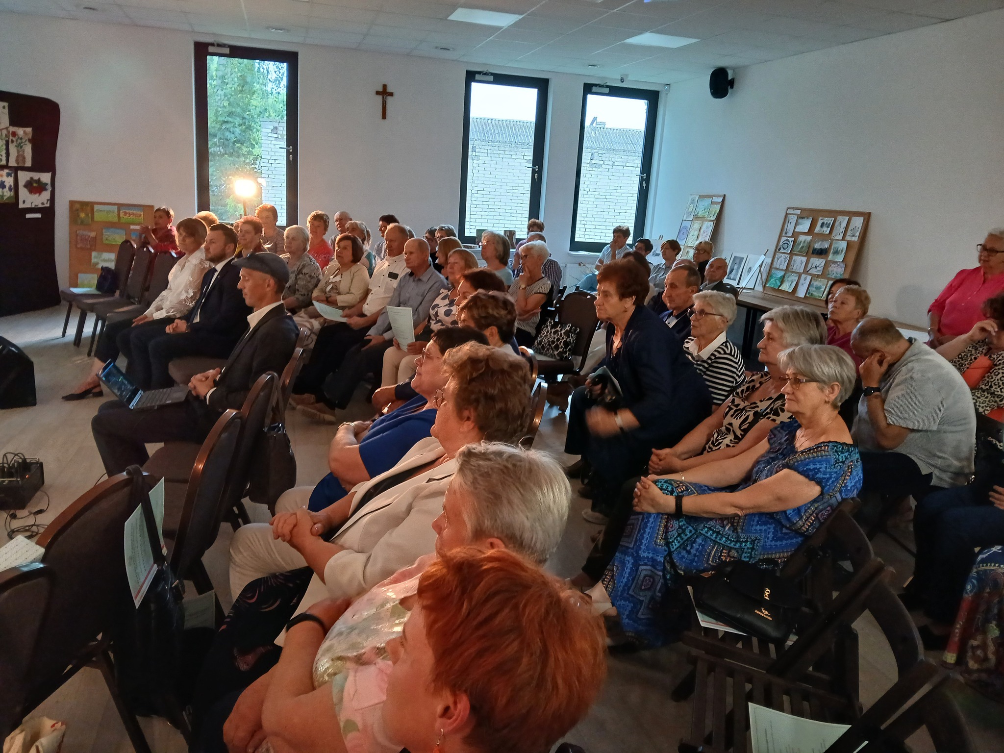 Grupa osób siedzi w pomieszczeniu z jasnymi ścianami, na białych krzesłach. Na ścianach wiszą obrazy i znajduje się krzyż. Okna wpuszczają naturalne światło.