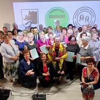 Grupa osób różnej wiekowo, uśmiechnięta, pozująca do zdjęcia w sali. W tle widoczne logo z połączeniem kolorów zieleni oraz bieli. Większość trzyma dokumenty.