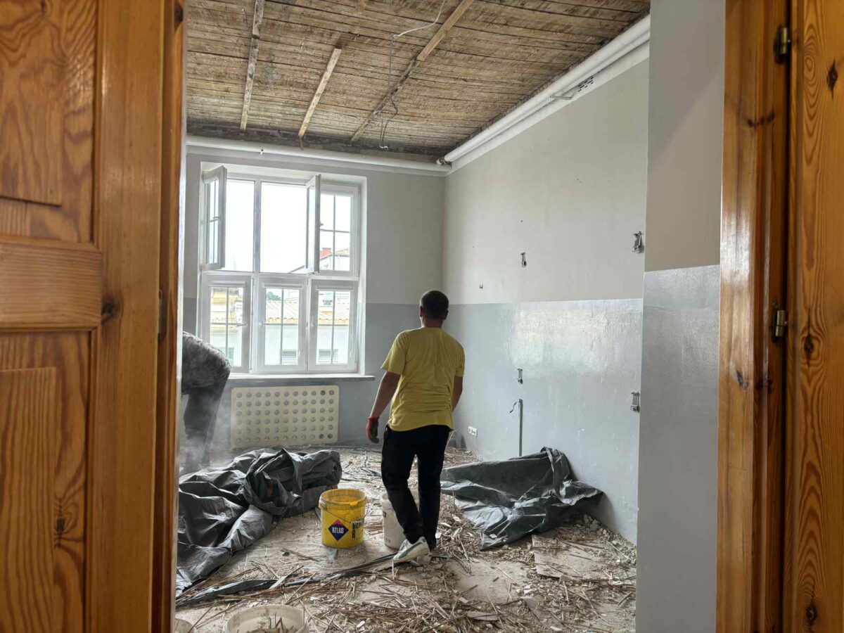 Pomieszczenie w remoncie z drewnianym sufitem i szarymi ścianami. Mężczyzna w żółtej koszulce i błękitne wiadro na podłodze. Duże okno wpuszcza światło dzienne.