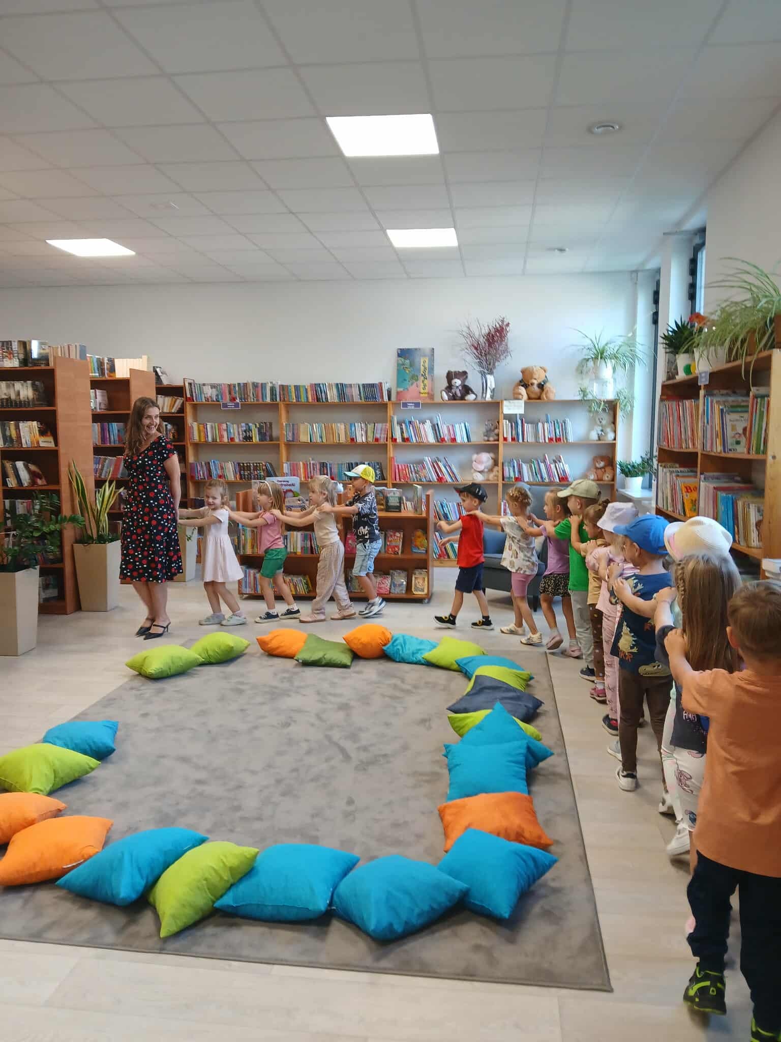 Grupa dzieci bawi się w sali z drewnianymi półkami pełnymi książek. Na podłodze leżą kolorowe poduszki. Ściany są jasne, a wnętrze jest dobrze oświetlone.
