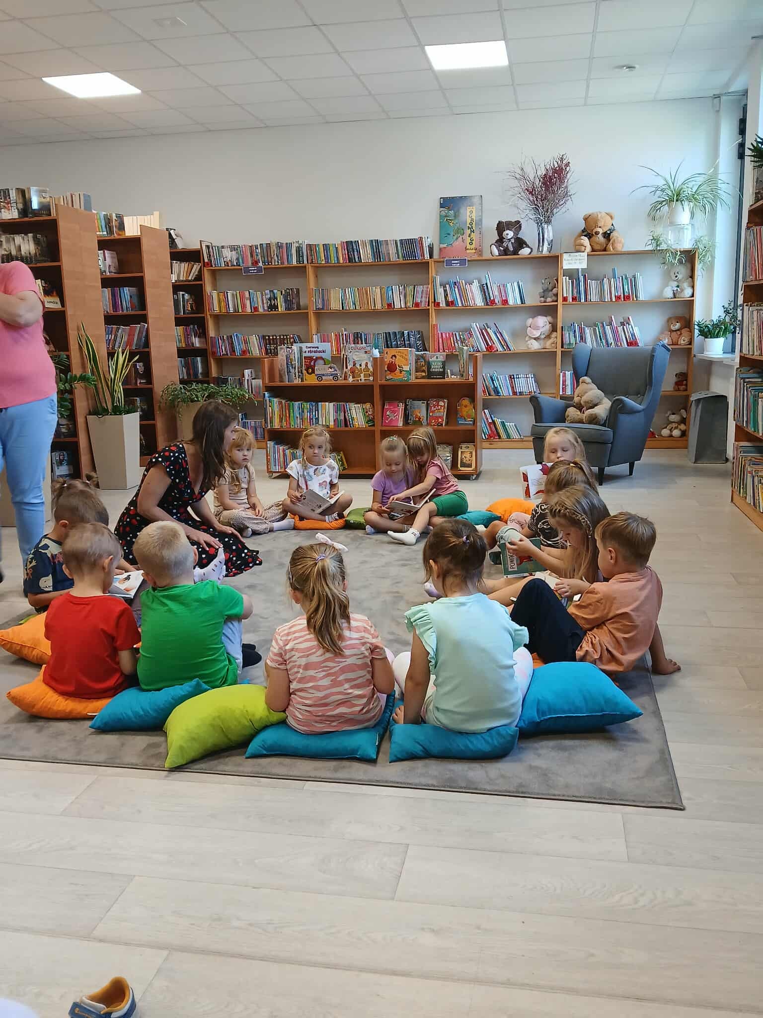 Dzieci siedzą na kolorowych poduszkach w bibliotece, zajęte książkami. Tło tworzą półki pełne książek, ściany jasne. Dookoła rośliny i maskotki dodają przytulności.