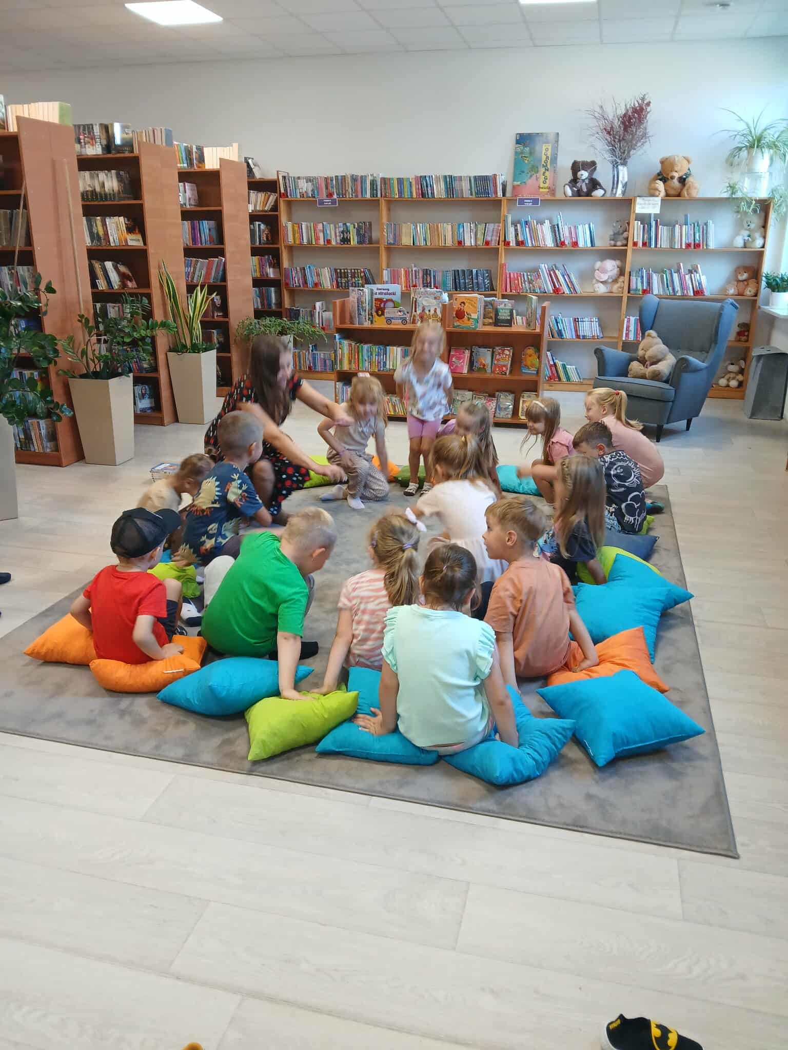 Dzieci siedzą na kolorowych poduszkach w jasnej bibliotece. Tło to półki z książkami i pluszowe misie. Rośliny dodają zieleni.