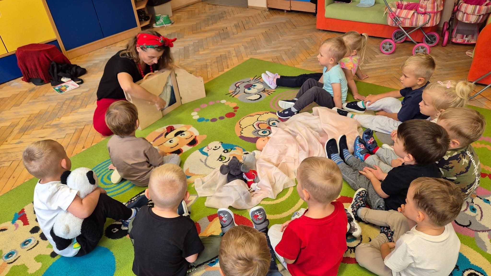 Dzieci siedzą w kręgu na kolorowym dywanie w przedszkolu. Kobieta z chustką na głowie pokazuje im pacynki. W tle są szafki i zabawki.