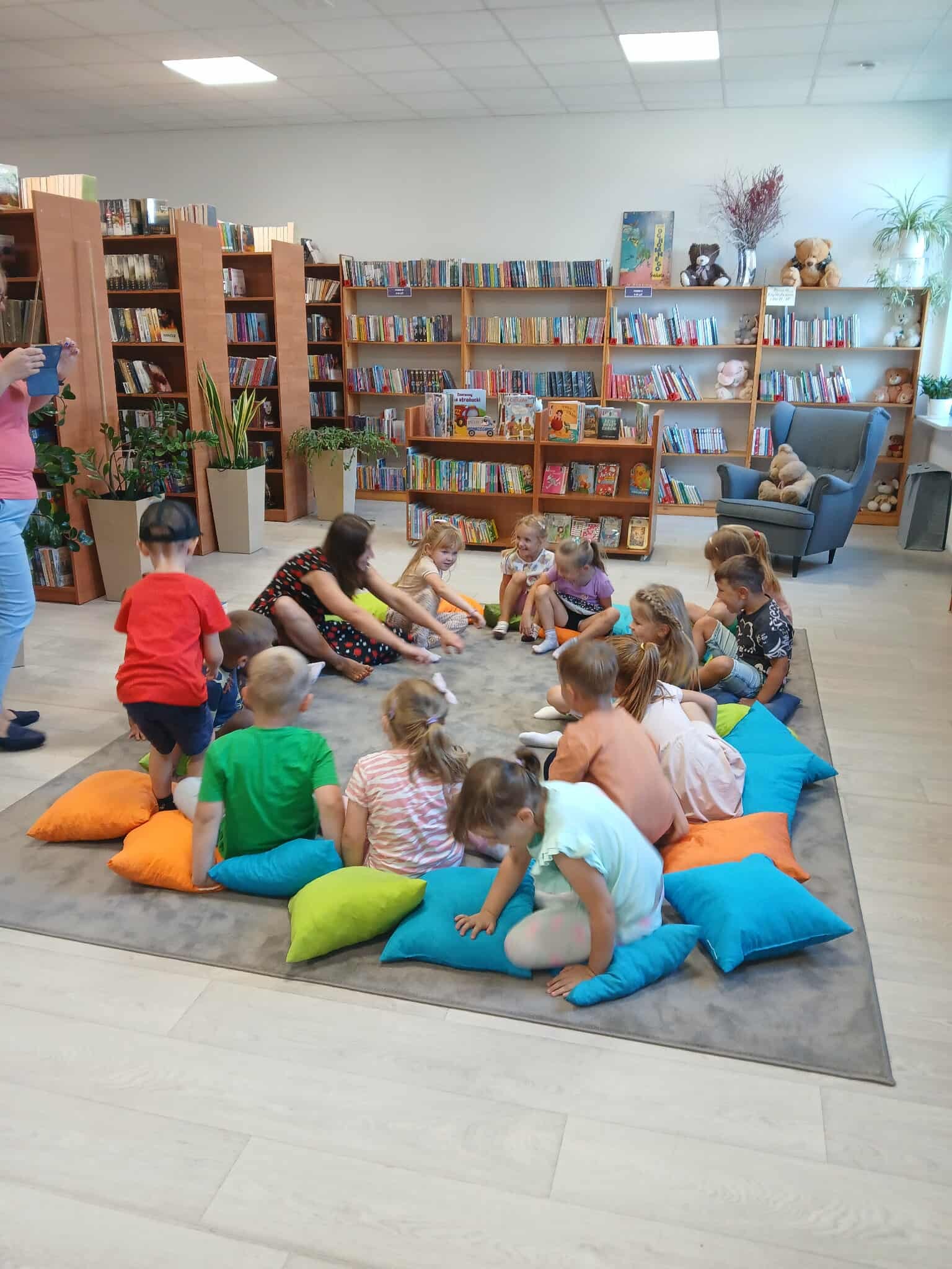 Grupa dzieci siedzi na kolorowych poduszkach w bibliotece. Na półkach w tle są książki i pluszaki. Oświetlenie jest jasne.