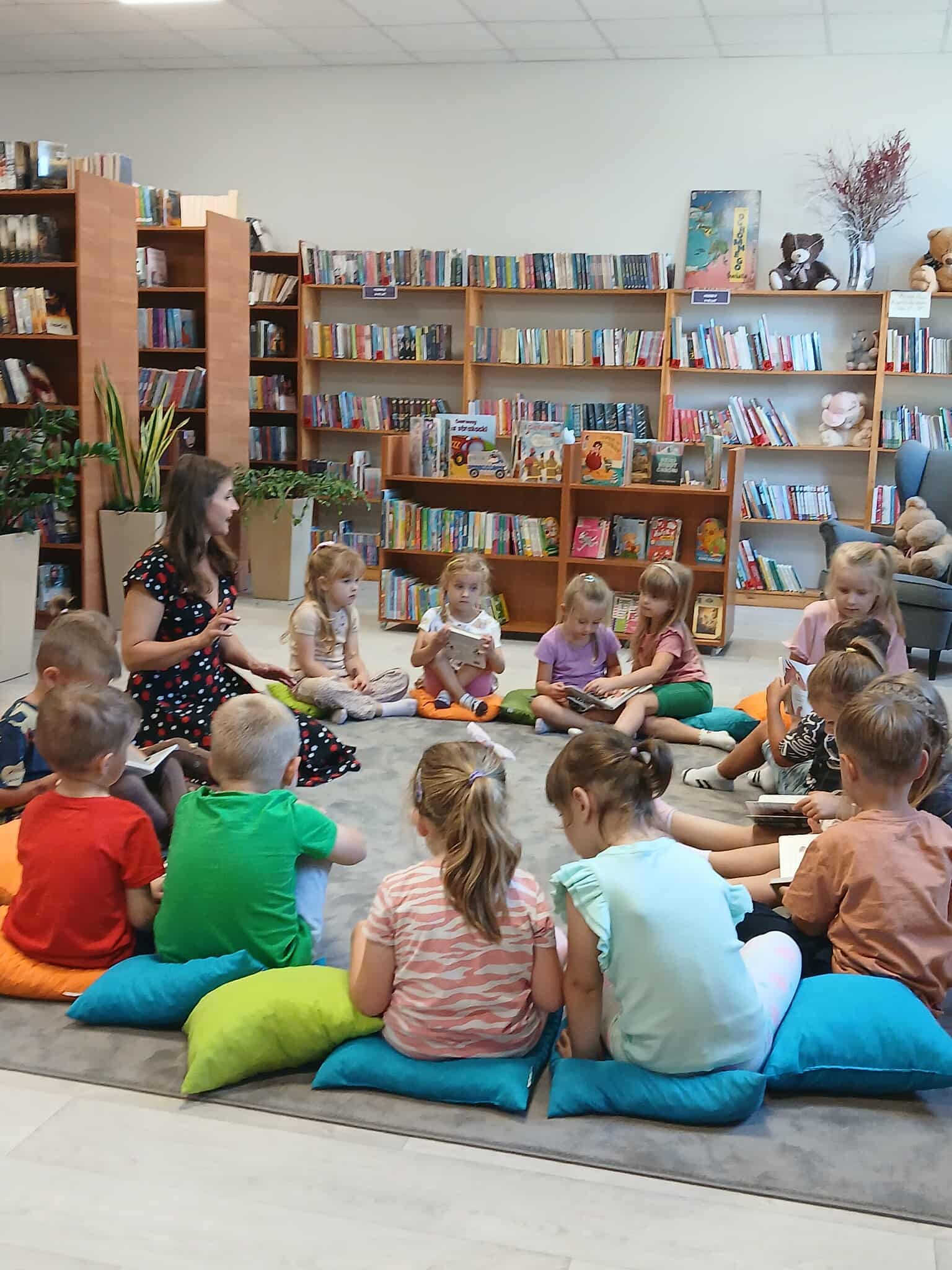 Grupa dzieci siedzi na kolorowych poduszkach w bibliotece. Przed nimi młoda kobieta czyta książkę. W tle regały z kolorowymi książkami.