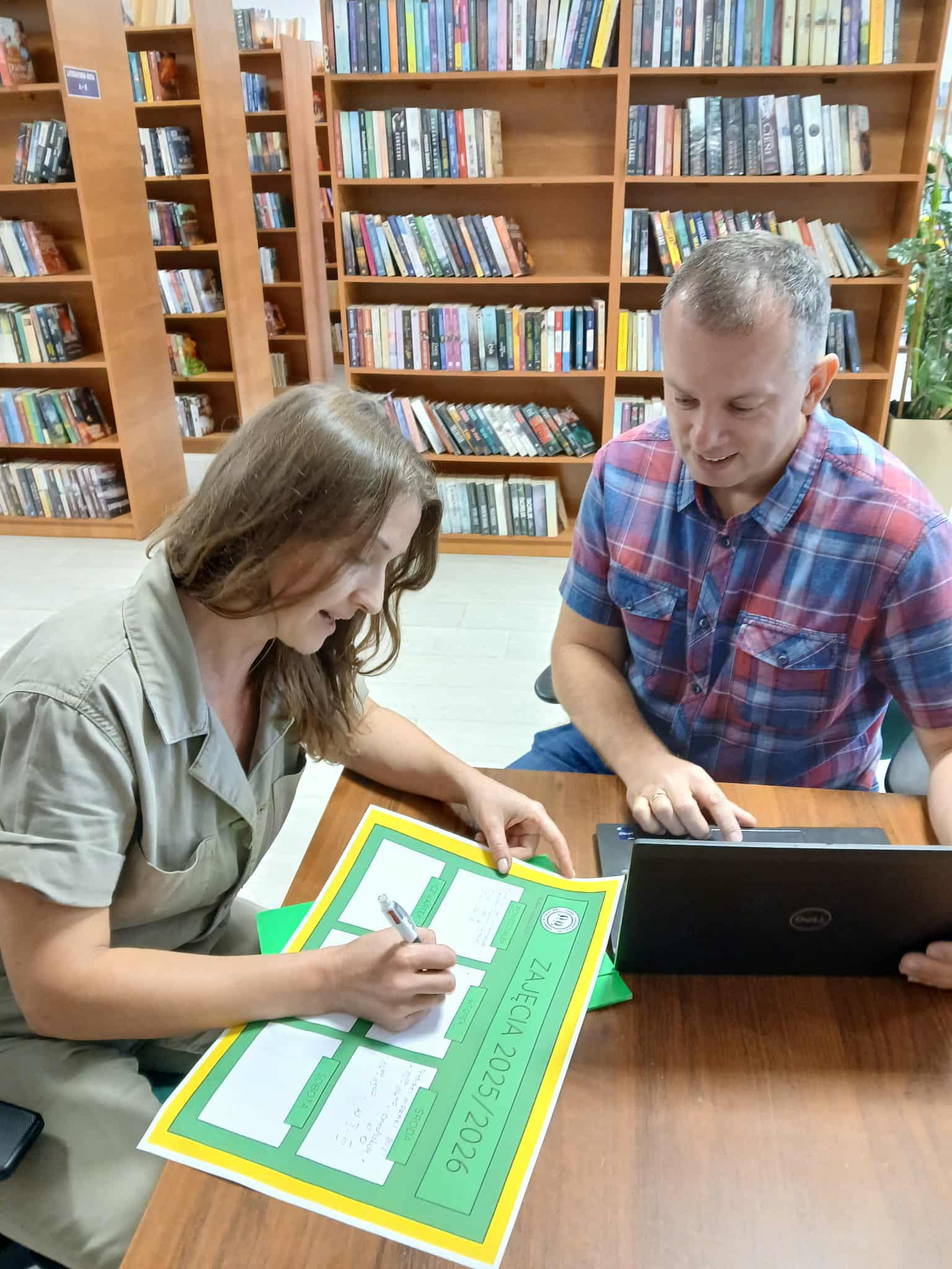 Dwoje ludzi pracuje przy stole w bibliotece. Kobieta pisze na zielono-żółtym formularzu. W tle widoczne są półki z książkami.