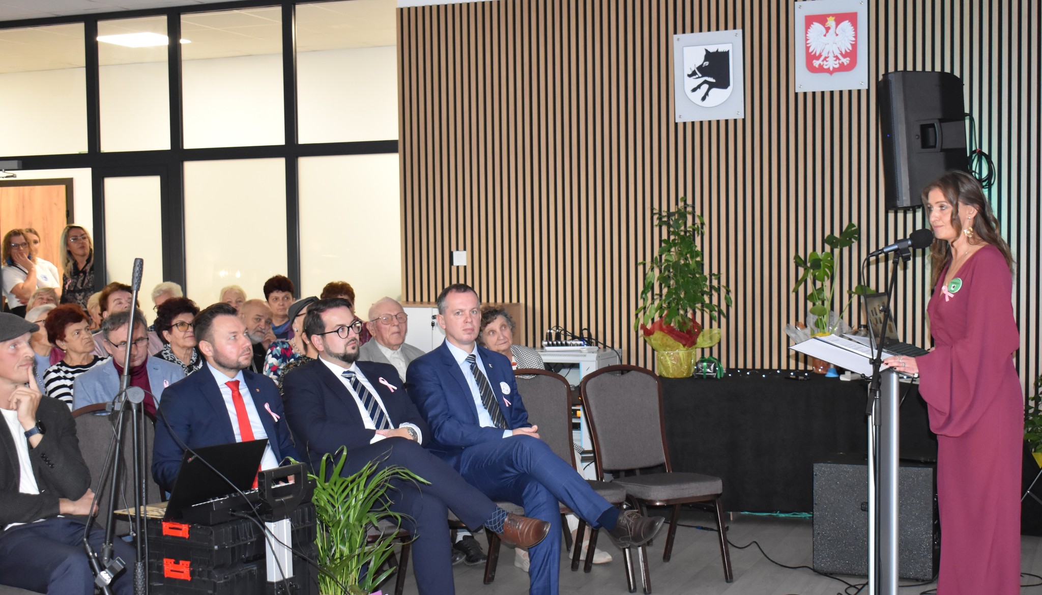 Grupa osób uczestniczy w spotkaniu w sali z drewnianymi panelami. Kobieta w bordowej sukience przemawia przy mikrofonie. W tle widać rośliny i symbole.