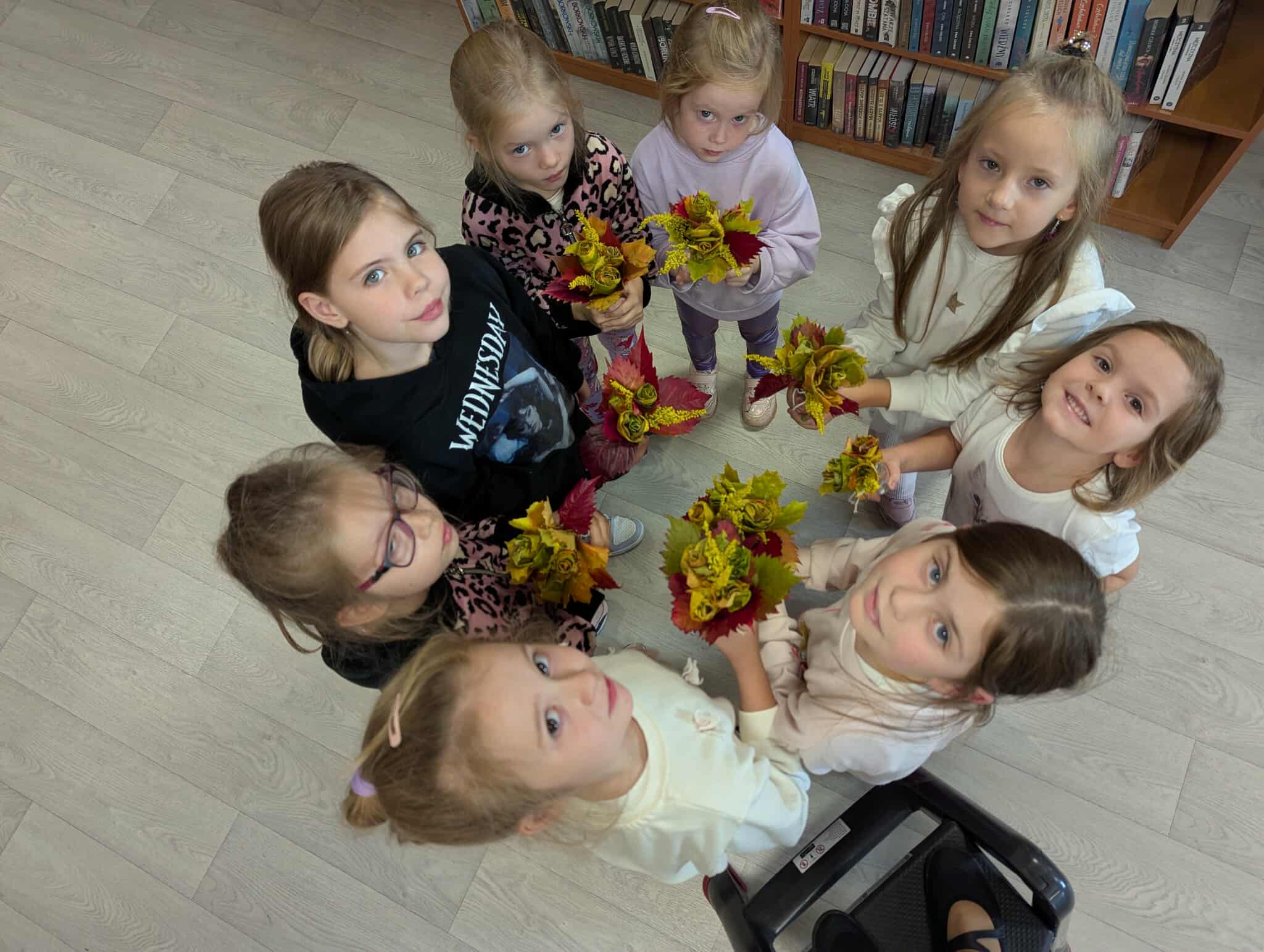 Grupa dzieci stoi na drewnianej podłodze, trzymając kolorowe bukiety. Dzieci są ubrane w jasne ubrania, a tło stanowi półka z książkami. Każde z nich patrzy w górę, do kamery.