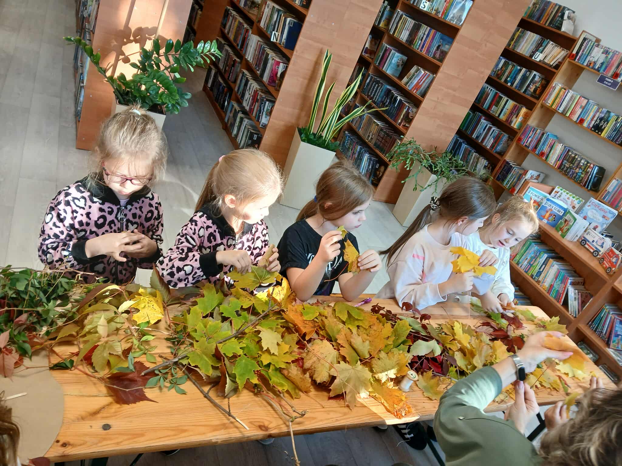 Dzieci przy stole pełnym kolorowych liści, w bibliotece z książkami na regałach. Słoneczne światło pada na wnętrze.