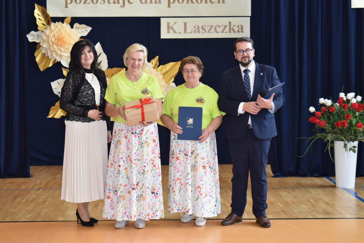 Cztery osoby stoją na tle ciemnoniebieskiej zasłony z dekoracją z papierowych kwiatów. Dwie kobiety w zielonych koszulkach i spódnicach w kwiaty trzymają nagrody. Obok stoją elegancko ubrani kobieta i mężczyzna.