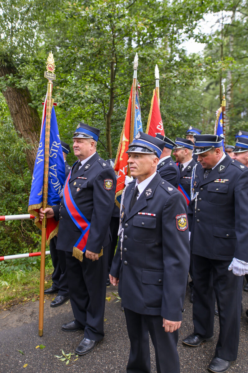 Grupa strażaków w mundurach z barwnymi sztandarami stoi na tle zielonego lasu. Dominuje kolor granatowy i czerwony. Tło jest pełne drzew i liści.