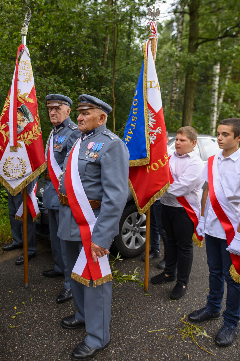 Na zdjęciu widać dwóch starszych mężczyzn w mundurach, z medalami, trzymających flagi. Tło stanowi zalesiony teren. Obok nich stoją młodsi chłopcy w białych koszulach z szarfami.