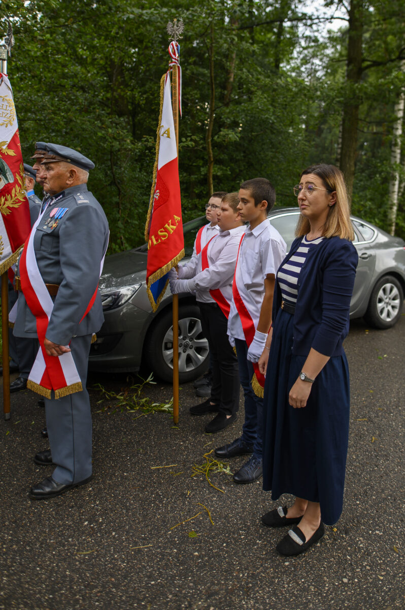 Grupa osób w odświętnych strojach stoi w szeregu z flagą Polski. Tło to zielony las. Przeważają kolory czerwony, biały i niebieski.