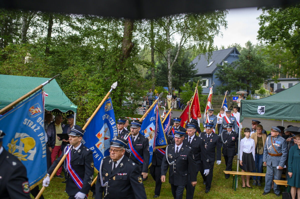 Parada strażaków w ciemnych mundurach maszerujących z niebieskimi i czerwonymi sztandarami. Tło: zielone drzewa, namioty, ludzie. Uroczystość pasuje do otoczenia w plenerze.