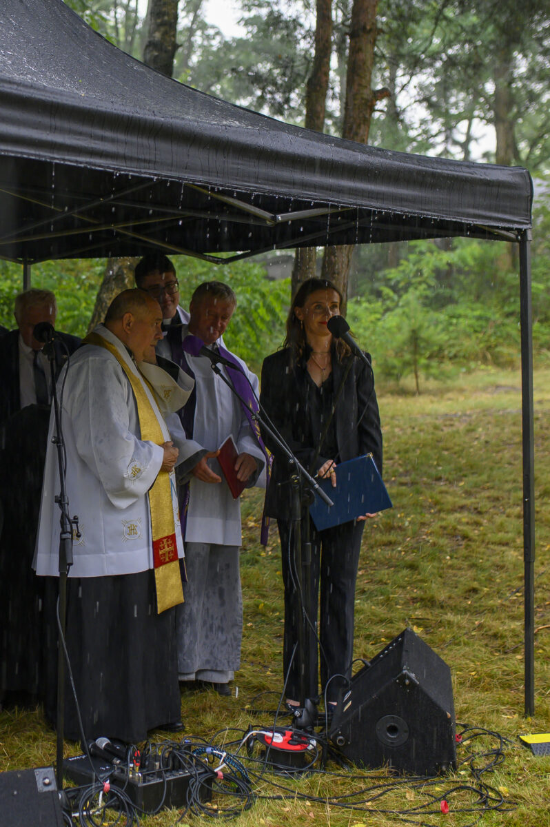Pod namiotem podczas deszczu stoi grupa osób w strojach liturgicznych i formalnych, mówiących do mikrofonów. Tło leśne.