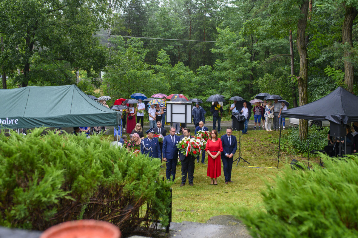 Zdjęcie przedstawia uroczystość na świeżym powietrzu. Grupa ludzi z wieńcami stoi na zielonej trawie. W tle są drzewa i namioty, a uczestnicy trzymają parasole.