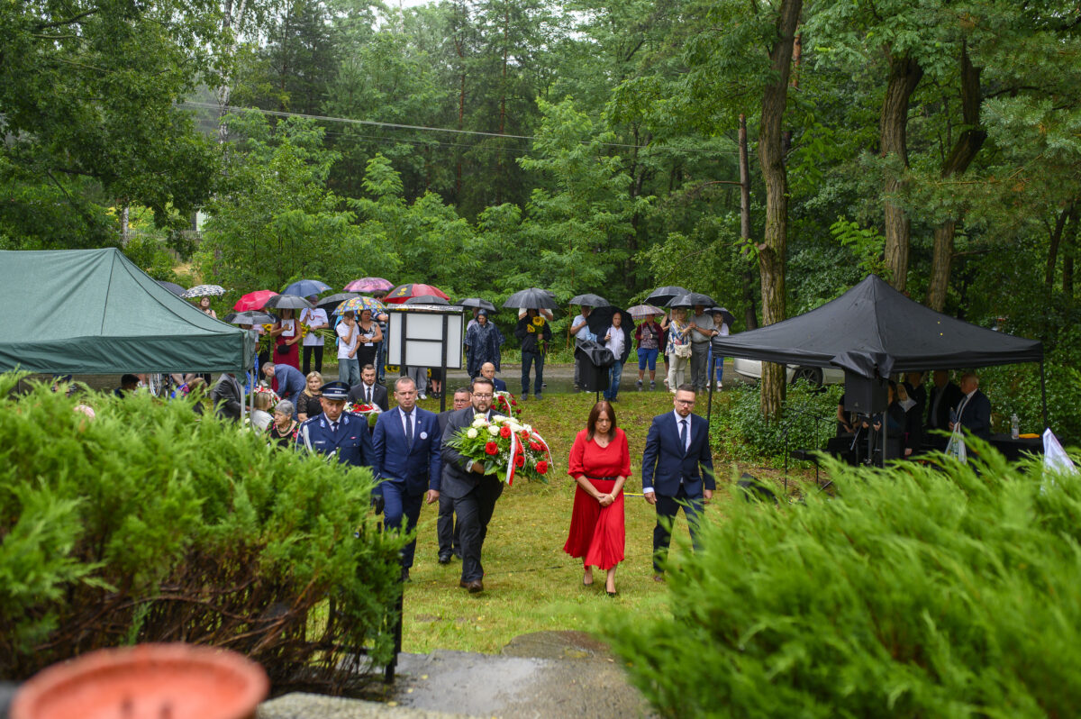 Grupa ludzi z kwiatami na pewno w uroczystości plenerowej. Zielona trawa, drzewa i namioty w tle. Parasole w różnych kolorach chronią przed deszczem.