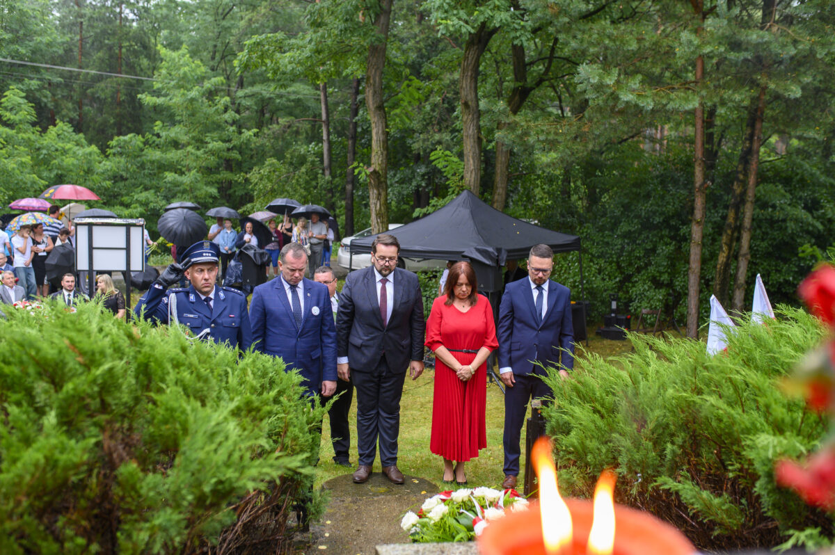 Pięć osób w eleganckich strojach stoi w lesie, obok złożonych kwiatów. Tło: zielone drzewa, ludzie z parasolkami. W tle czarny namiot.