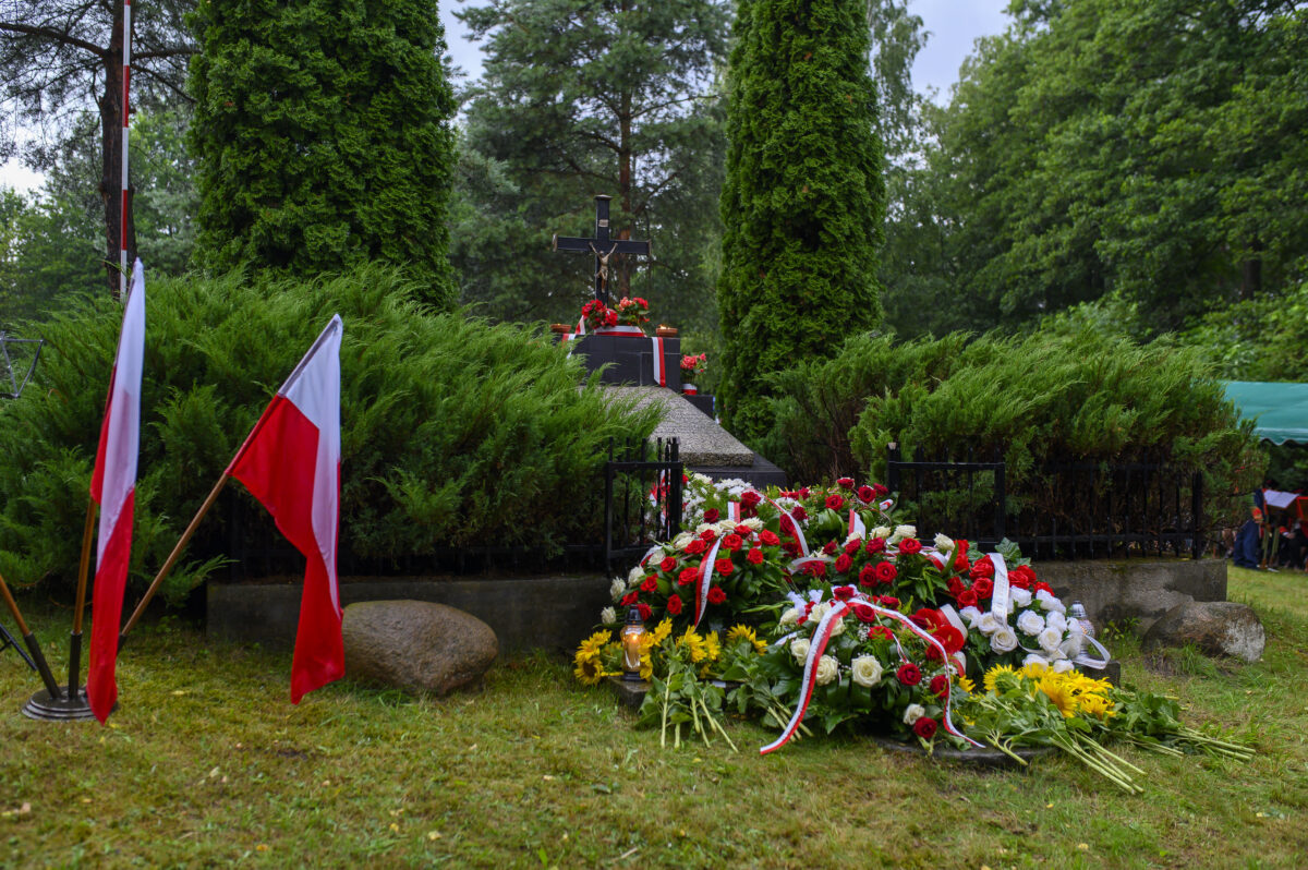 Nagrobek z krzyżem otoczony zielenią, kwiaty biało-czerwone. Polska flaga z lewej, tło to drzewa i trawa.