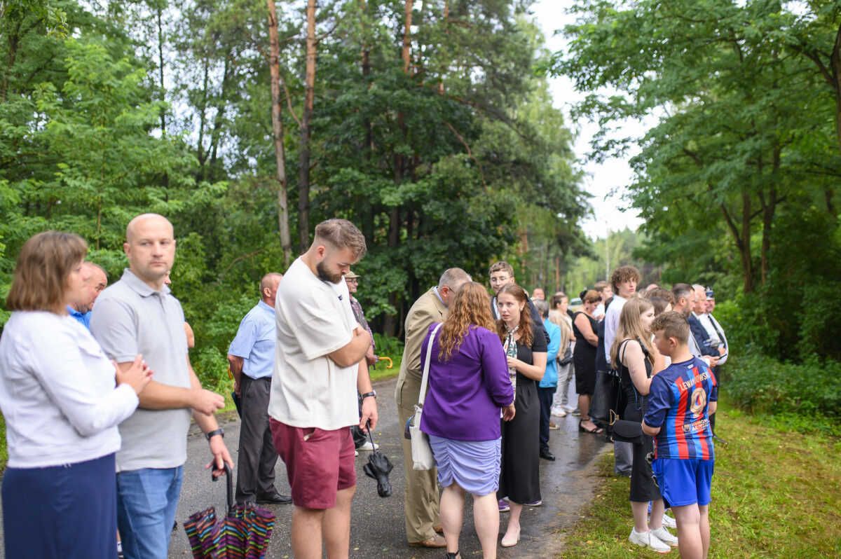 Grupa ludzi stoi na leśnej ścieżce, otoczona zielenią drzew. Ubrani są w różnorodne stroje od casualowych po eleganckie. Tło stanowią bujne drzewa i roślinność.