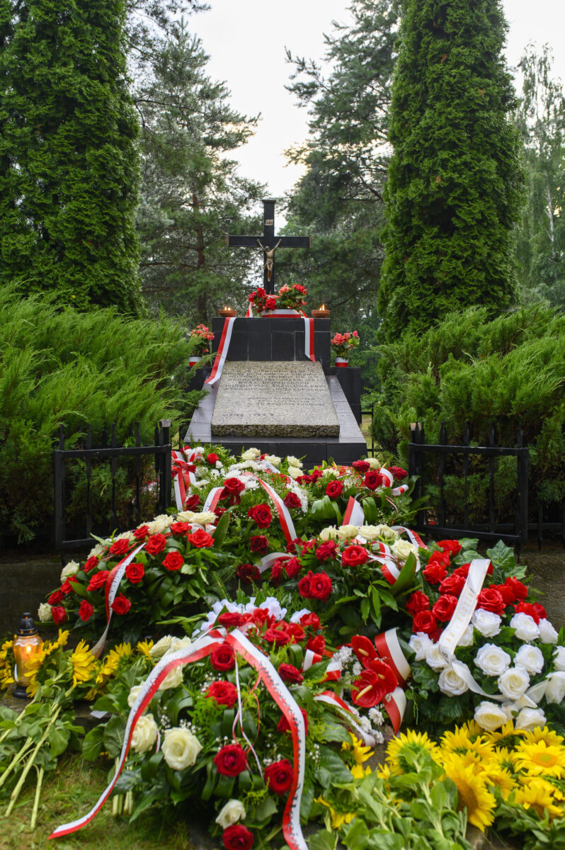 Na zdjęciu widoczny jest grób ozdobiony wieńcami z czerwonych i białych kwiatów, z krzyżem w tle. Tło to zielone drzewka i krzewy. Na wieńcach widoczne są biało-czerwone wstęgi.