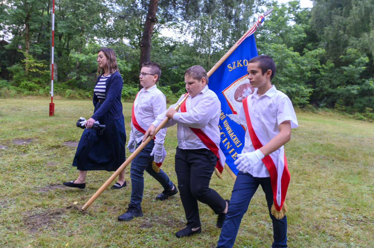 Na zdjęciu grupa młodych osób w białych koszulach niesie niebieską flagę. Tło stanowi zielona trawa i drzewa. Atmosfera jest uroczysta, a kolory żywe.