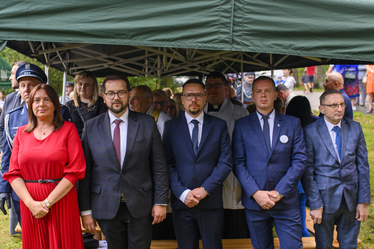 Pod zielonym namiotem stoi grupa elegancko ubranych osób. Mężczyźni w garniturach, kobieta w czerwonej sukience. W tle widać uczestników wydarzenia.