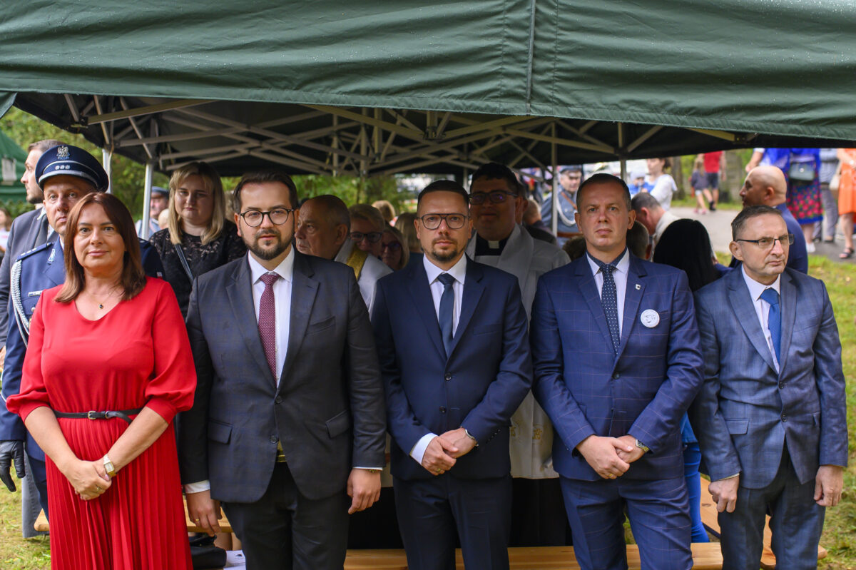 Grupa ludzi w eleganckich strojach stoi pod zielonym namiotem. Mężczyźni w garniturach, kobieta w czerwonej sukience. W tle widać osoby i zieleń.