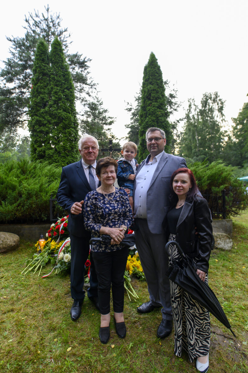 Grupa pięciu osób stoi na tle zielonych drzew i krzewów. Mężczyźni mają na sobie garnitury, a kobiety eleganckie ubrania. Widać kwiaty i znicze na trawie.