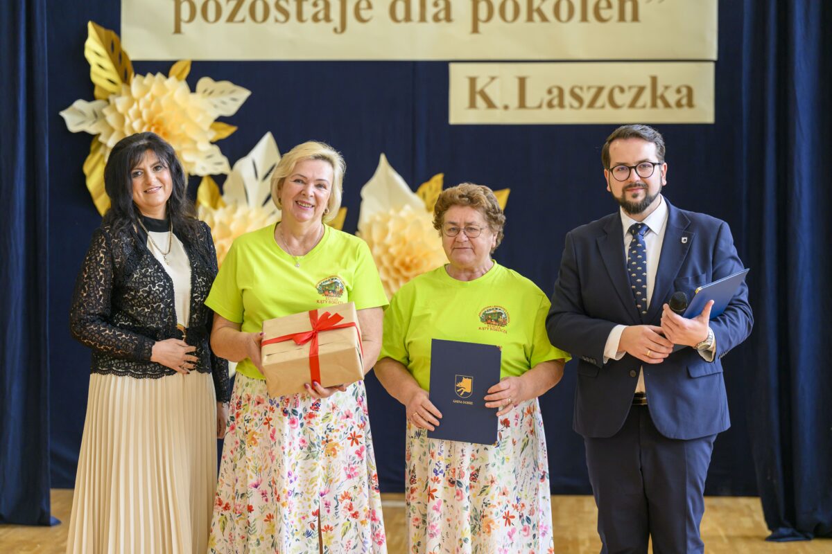 Na zdjęciu cztery osoby na tle dekoracji z żółtych i białych kwiatów. Dwie kobiety w zielonych koszulkach trzymają prezenty. Mężczyzna i kobieta są elegancko ubrani.